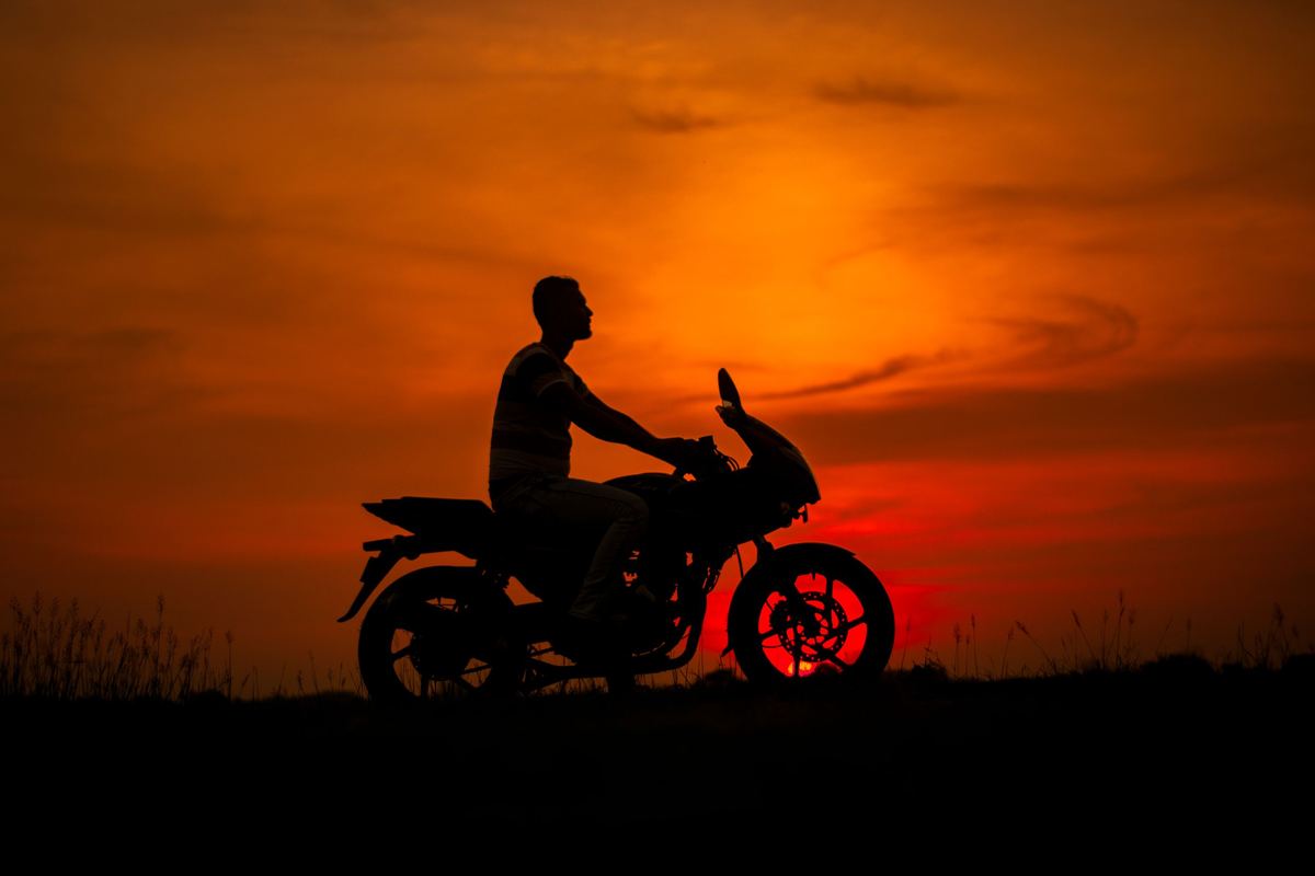 Silueta de un motociclista sobre su moto recortada contra un cielo naranja al atardecer.